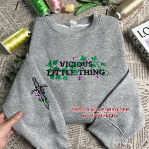 Pu&ograve; includere: Felpa grigia con il testo ricamato "Vicious Little Thing" e un pugnale con fiori. Il testo &egrave; in nero con viti verdi e fiori viola. Il design &egrave; ricamato sul davanti della felpa. Il testo "Design and Embroider by HSTHreads" &egrave; stampato sotto il design.
