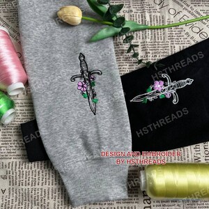 Pu&ograve; includere: Due disegni ricamati di un pugnale con fiori. Il pugnale &egrave; bianco con contorni neri e i fiori sono viola. I disegni sono su tessuto grigio e nero. Il testo "DESIGN AND EMBROIDER BY HSTHREADS" &egrave; sotto i disegni.