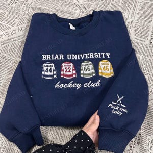 Könnte beinhalten: Marineblaues Briar University Hockey Club Sweatshirt. Mit gestickten Hockey-Trikots mit Namen und Nummern. Der Ärmel hat den Text "Puck me, baby" mit gekreuzten Hockeyschlägern. Ein bequemes, lässiges Oberteil.