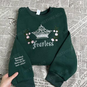 Puede incluir: Una sudadera de color verde oscuro con una corona blanca y flores rosas bordadas en la parte delantera. El texto "Fearless" está bordado debajo de la corona. La sudadera tiene mangas largas y cuello redondo.