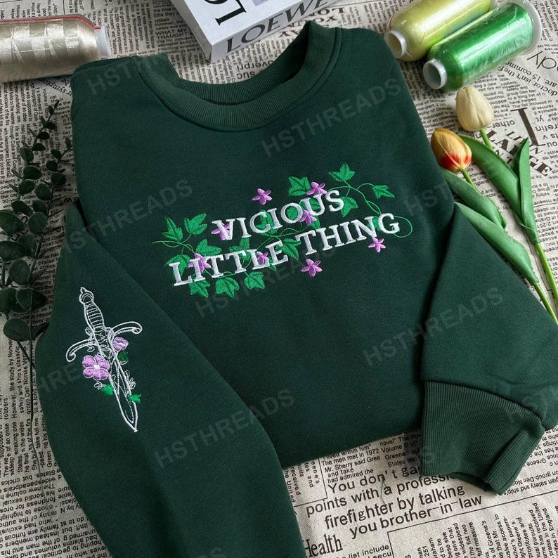 Pu&ograve; includere: Una felpa verde scuro con il testo "VICIOUS LITTLE THING" ricamato in bianco con fiori viola. Un pugnale bianco con fiori viola &egrave; ricamato sulla manica sinistra.