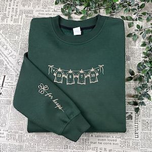 May include: A dark green crewneck sweatshirt featuring an embroidered design of four jerseys hanging on a clothesline with the numbers 13, 6, 7, and 10. The sleeve displays an embroidered four-leaf clover and the cursive text for keeps.