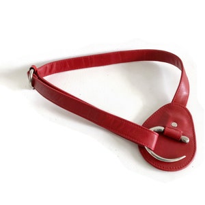 May include: A red leather belt with a silver buckle. The buckle is a large, oval-shaped piece of metal with a curved opening.