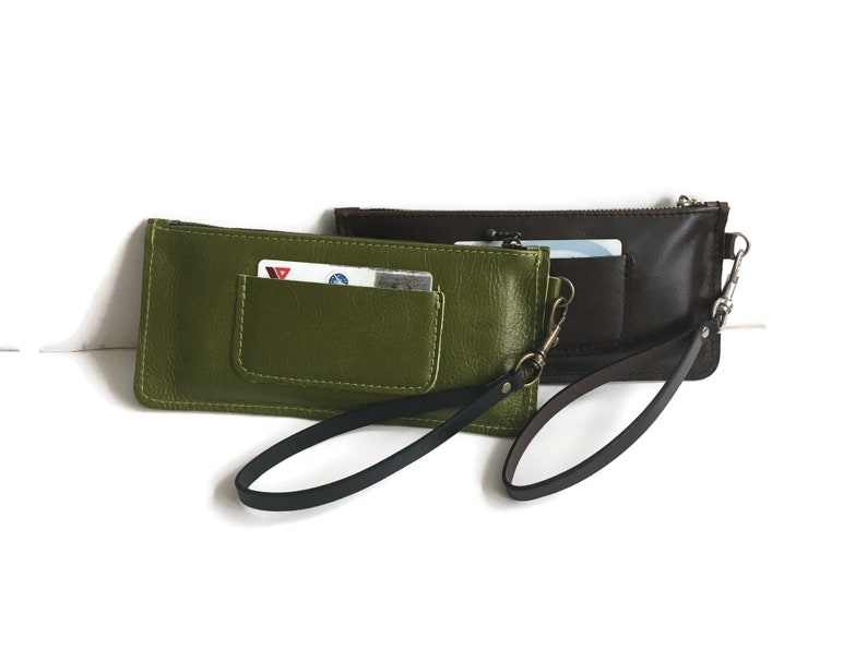 May include: Two leather wallets with wrist straps. One is green and the other is brown. Both have a zipper closure and a small pocket on the front.