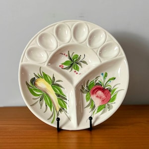 May include: A white ceramic serving platter with a divided design. The platter features a section for eggs and three sections decorated with hand-painted vegetables in green, yellow, and red. The platter is displayed on a black metal stand.
