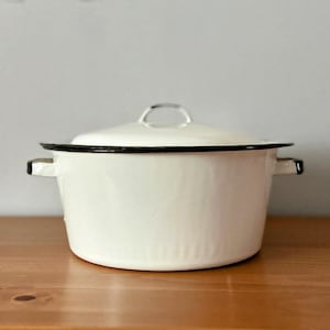 May include: A white enamel pot with a matching lid, featuring a black rim around the top edge and the lid. The pot has two side handles and a small metal handle on the centre of the lid. The finish is smooth and glossy with a rounded shape.