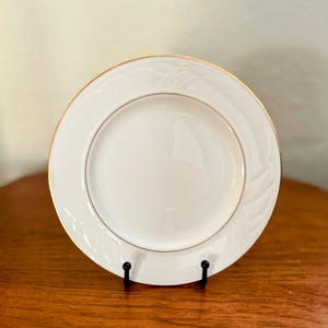 May include: A white ceramic plate with a gold rim and a raised floral design. The plate is on a wooden surface.