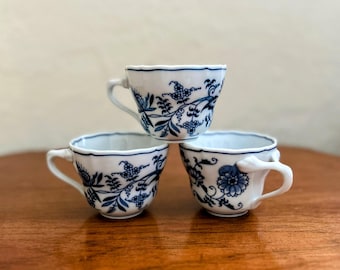 May include: Three white teacups with blue floral patterns stacked on top of each other. The cups have a scalloped rim and a delicate floral design.