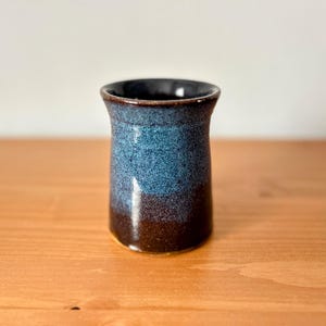 May include: A handmade ceramic vase with a speckled blue and brown glaze. The vase has a cylindrical shape with a slightly flared rim. The interior is dark, and the exterior features a gradient of colors.