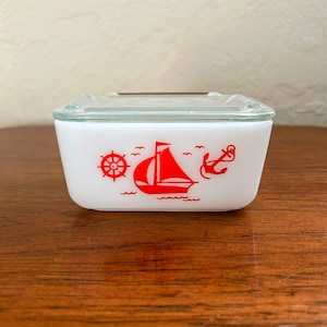 Puede incluir: Contenedor de almacenamiento de vidrio rectangular blanco con una tapa transparente. El contenedor tiene un diseño náutico rojo con un velero, un ancla y un timón.