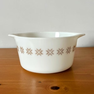 Op de afbeelding: Witte keramische kom met bruin sterrenpatroon rond de rand. De kom heeft twee handvatten en staat op een houten oppervlak. Een vintage keukenartikel.