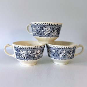 May include: Three cream-colored teacups with a blue and white floral design. The cups have a repeating pattern of leaves and berries around the rim. Each cup has a curved handle.
