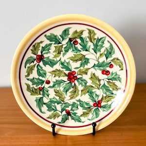 May include: A round decorative plate with a festive holly berry design. The plate features a cream-colored center with green leaves and red berries. A thin red band and a yellow border frame the design. The plate is displayed on a black stand.