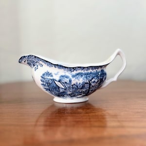 May include: A white ceramic gravy boat with a blue and white floral design. The boat has a wide, rounded base and a spout on one side.