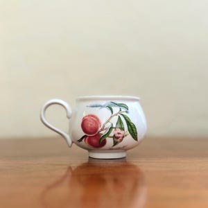 Może przedstawiać: Biały ceramiczny kubek do herbaty z wzorem brzoskwini. Kubek ma zaokrąglone ciało i uchwyt. Wzór przedstawia gałąź brzoskwiń z liśćmi i kwiatami.