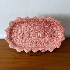 Könnte beinhalten: Rosa Keramik-Serviertablett mit einem dekorativen Blumen- und Hasen-Design. Das Tablett hat einen gewellten Rand mit geprägten Rosen und zwei kreisförmigen Abschnitten mit Reliefbildern von Hasen. Das Tablett wird von einem goldenen Metallständer getragen.
