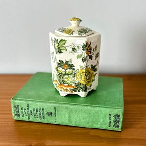 May include: A hexagonal ceramic jar with a lid, featuring a floral design in yellow, green, and brown on a cream background. The jar is placed on a green book with text. The jar is approximately 5 inches tall.