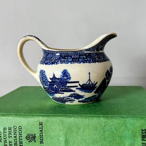 Puede incluir: Un pequeño creamer blanco con un patrón de sauce azul y blanco. El creamer tiene un cuerpo redondeado y un pequeño pico. Está sentado encima de un libro verde.