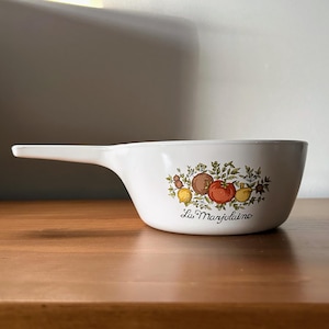 Może przedstawiać: Biały ceramiczny garnek z długim uchwytem i kwiatowym wzorem przedstawiającym pomidory, cytryny i zioła. Tekst "La Marjolaine" jest nadrukowany na spodzie garnka.