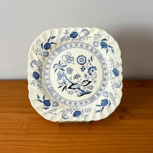 Könnte beinhalten: Quadratischer, cremefarbener Teller mit blauem Blumendesign. Der Teller hat einen gewellten Rand und ein zentrales kreisförmiges Muster mit Blumen und Blättern. Blaue Blumenmotive befinden sich ebenfalls am Rand.