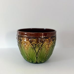 Puede incluir: Un cuenco de cerámica con borde e interior marrón oscuro. El exterior presenta un diseño floral verde y beige. El cuenco tiene un acabado brillante y probablemente se utiliza con fines decorativos o como maceta.