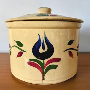 Puede incluir: Un tarro de cerámica de color crema con tapa, que presenta un diseño floral pintado a mano en azul, verde y rojo. El tarro tiene una forma redonda y una superficie lisa.