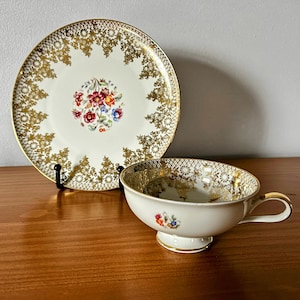 May include: A vintage teacup and saucer set with a white background and gold floral pattern. The cup and saucer have a delicate gold trim.