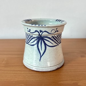 May include: A ceramic vase with a light blue and white speckled finish. The vase features a dark blue leaf and lattice design around the middle and a dark blue rim. The vase is approximately 5 inches tall.