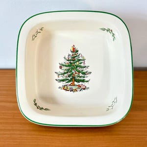 May include: Square, off-white ceramic serving dish with a green border. The center features a detailed illustration of a decorated Christmas tree with presents at the base. Small sprigs of greenery are in each corner.