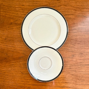 Puede incluir: Dos platos de cerámica blanca con borde negro. El plato más grande es un plato para cenar y el plato más pequeño es un platillo.