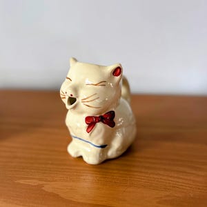 Op de afbeelding: Een crèmekleurige keramische kattenmelkkan met een rode vlinderdas en rode accenten op de oren. De kat heeft gesloten ogen en een schenktuit. Een blauwe lijn accentueert de basis. Vintage stijl.