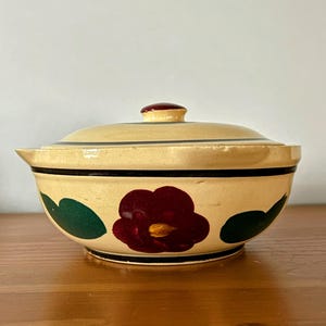 Peut inclure: Un bol en céramique couvert, orné d'un motif floral. Le bol est de couleur crème avec une fleur rouge foncé et des feuilles vertes peintes sur le côté. Une bande noire entoure le bol, et le couvercle a un petit bouton rouge.