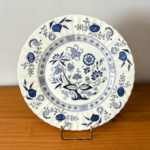 May include: A white ceramic plate with a blue floral design. The plate has a scalloped edge and a central floral motif with intricate details. The blue and white color scheme gives the plate a classic, vintage feel. The plate is displayed on a gold-colored stand.