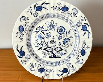 May include: A white ceramic plate with a blue floral design. The plate has a scalloped edge and a central floral motif with intricate details. The blue and white color scheme gives the plate a classic, vintage feel. The plate is displayed on a gold-colored stand.