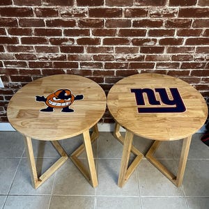 Op de afbeelding: Twee houten bijzettafels met ronde bladen. De ene tafel heeft een sticker met een cartoonfiguur en de andere heeft een sticker met het logo van de New York Giants.
