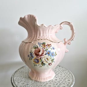 May include: A pink ceramic pitcher with a floral design. The pitcher has a ruffled top and a gold trim around the rim. The floral design features pink, yellow, blue, and green flowers.