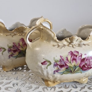 May include: Two antique porcelain flower pots with ornate handles and floral designs. The pots are cream-coloured with pink and gold accents.
