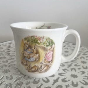 May include: White ceramic mug with a charming illustration of rabbits in a garden. The design features rabbits in blue and pink outfits. The mug has a curved handle and a slightly flared rim.