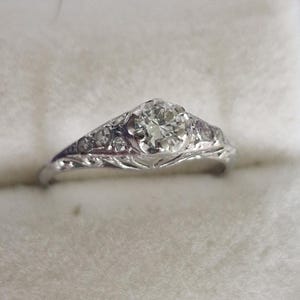 May include: A silver-coloured metal ring featuring a central round-cut clear gemstone set in a prong setting. The band is decorated with intricate filigree metalwork and smaller clear accent stones embedded along the shoulders of the ring shank.