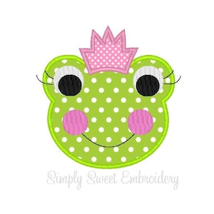May include: Green frog face with pink polka dots and a pink crown. The frog has large black eyes with eyelashes and pink cheeks.