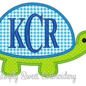 Può includere: Una tartaruga ricamata verde e blu con un guscio a quadri blu e le lettere "KCR" in blu sul guscio.