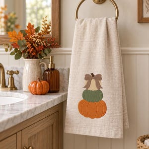 May include: A beige hand towel featuring an embroidered stack of three pumpkins in orange, green, and cream, topped with a brown bow. The towel is hanging from a circular brass ring fixture, displaying a textured fabric weave throughout the design.