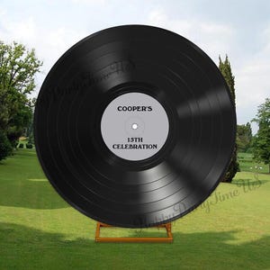 May include: A large black vinyl record with the text "COOPER'S 15TH CELEBRATION" in white. The record is displayed on a wooden stand on a grassy lawn, with trees and a cloudy sky in the background.