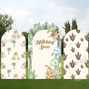 May include: Three arched backdrop panels featuring a dinosaur theme. The left panel shows volcanoes and palm trees. The centre panel displays the text Hatching Soon with illustrations of dinosaurs and tropical leaves. The right panel features dinosaur footprints.