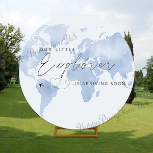 May include: A round, white sign with a light blue world map design. The text "Our Little Explorer is Arriving Soon" is written in elegant script. A small airplane and heart graphic are included. The sign is displayed on a wooden stand.
