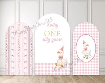 May include: Three arched party backdrops with pink and white designs. One features a bow pattern, another has text that reads "Kolby is ONE silly goose", and the third has a goose illustration. "Happy Party Time Us" is written on the backdrops.