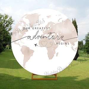 Könnte beinhalten: Rundes Schild mit Weltkarten-Design in neutralen Tönen, mit dem Text "Our Greatest Adventure Begins". Eine kleine Flugzeuggrafik und eine herzförmige gepunktete Linie sind ebenfalls enthalten. Das Schild wird auf einem Holzständer in einer grasbewachsenen Umgebung im Freien präsentiert.