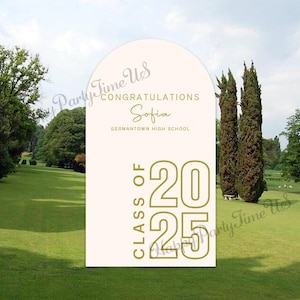 May include: A white arch with gold text that reads "Congratulations Sofia Germantown High School Class of 2025". The arch is in front of a green grassy field with trees in the background.
