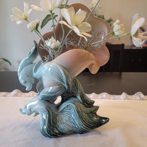 May include: A ceramic flower vase with a unique design, featuring a light blue base and a peach-colored top. White flowers are arranged in the vase. The vase has a glossy finish and a sculptural, organic form.
