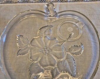 May include: Clear glass apple-shaped serving dish with a raised floral design. The dish features a stem handle and a detailed embossed flower and leaf pattern. The clear glass allows the design to be visible from both sides, making it a decorative and functional piece.